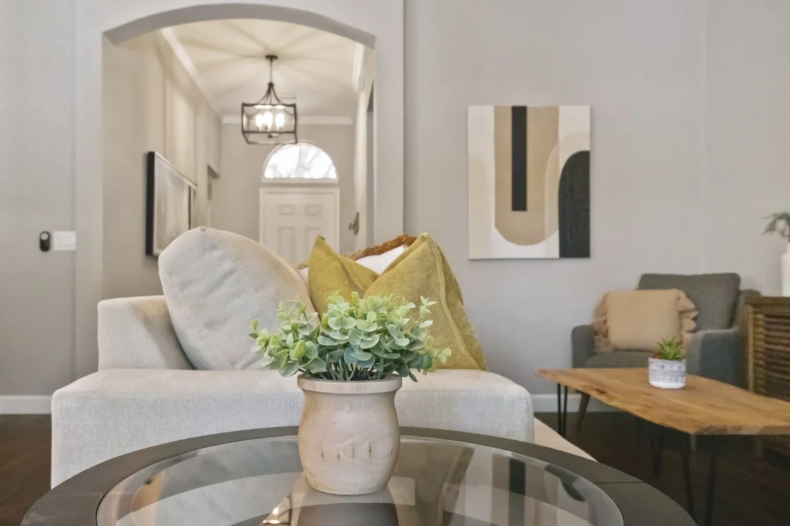 A vase of plants adorns a glass table in the living room, offering a view of the hallway that leads to the front door.