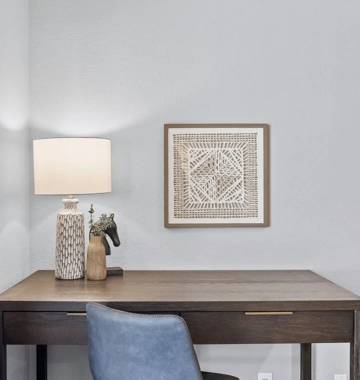 A workspace in the corner of the bedroom includes a desk, a lamp, a picture on the wall, and a cushy gray velvet office chair.