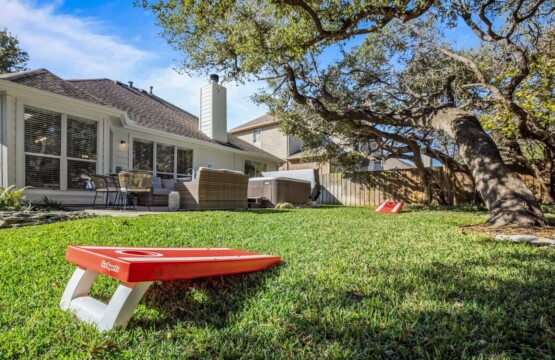 A spacious backyard features lush green grass and trees, complemented by a patio with a hot tub. The house, with its wide glass windows, serves as a backdrop, while a rustic wooden fence adds to the charm.