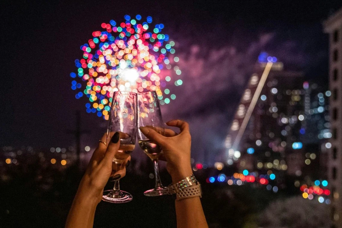 A couple's hand raising a toast with new year's eve fireworks and the city of cedar park at the backdrop