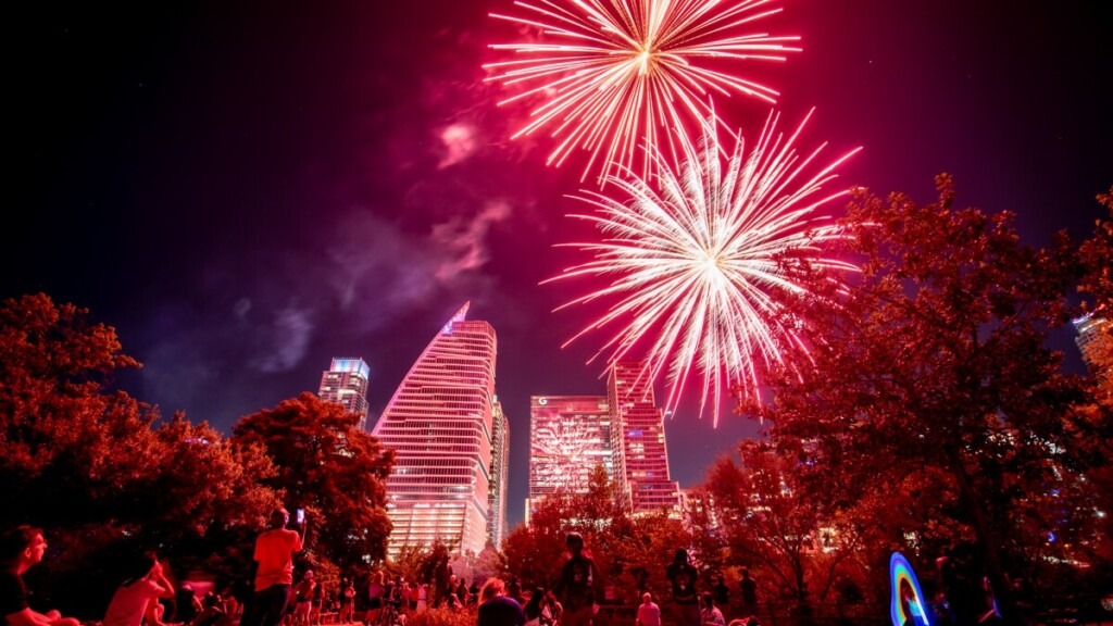 Fireworks display lighting up the night sky during the Cedar Park 4th of July Celebration, with vibrant red and pink bursts over a festive crowd gathered between Cedar Park and Austin, Texas.