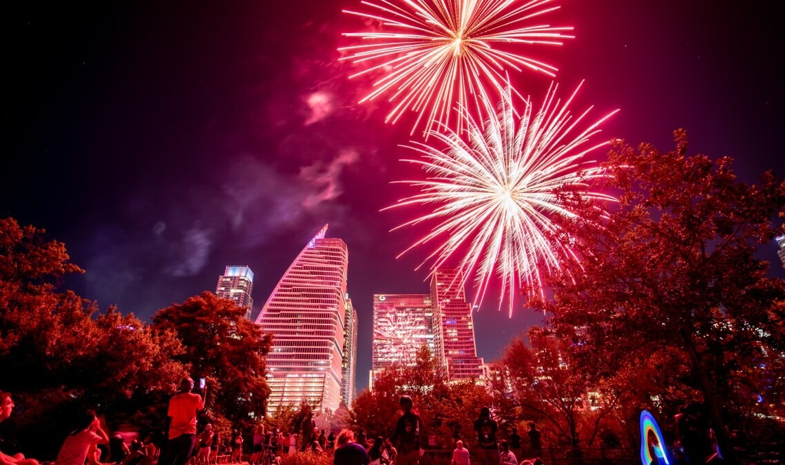 Fireworks display lighting up the night sky during the Cedar Park 4th of July Celebration, with vibrant red and pink bursts over a festive crowd gathered between Cedar Park and Austin, Texas.