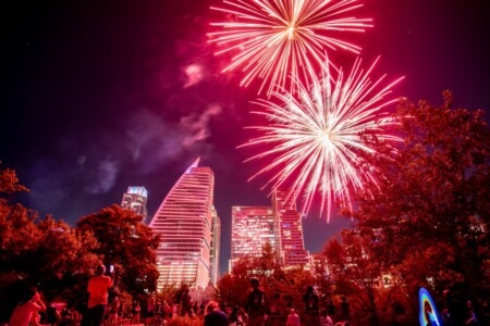 Fireworks display lighting up the night sky during the Cedar Park 4th of July Celebration, with vibrant red and pink bursts over a festive crowd gathered between Cedar Park and Austin, Texas.