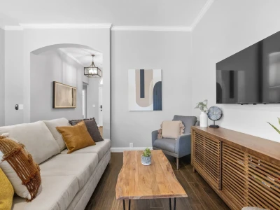 A stylish and charming living room features a cushy couch with pillows, centered around a low coffee table. To the right, a wall-mounted smart TV is paired with a wooden low cabinet, while a single cushy chair sits in the corner.