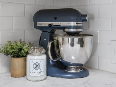 A blue mixer with a stainless steel bowl sits beside a candle and a small potted plant in the corner of the kitchen counter. Electrical sockets are located on the right side.