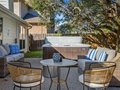 A patio features a hot tub paired with rattan couches adorned with cushions and pillows, alongside a round table and lawn chairs. This cozy space is nestled beneath tree branches and framed by a wooden fence.