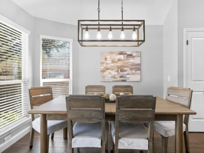 A rustic dining table with six chairs sits beneath a chandelier, with large windows on the left offering a full view of the backyard.