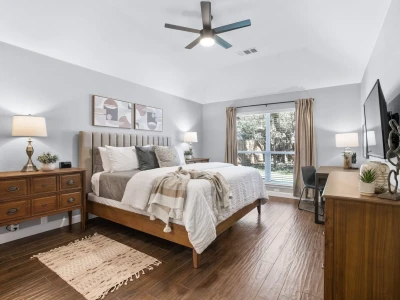 A spacious bedroom with hardwood floors features a king-size bed with a plush mattress, pillows, and linens, flanked by tables with lamps on both sides. In the corner, there's a work desk, along with a flat-screen smart TV, a ceiling fan, and a view of the backyard.